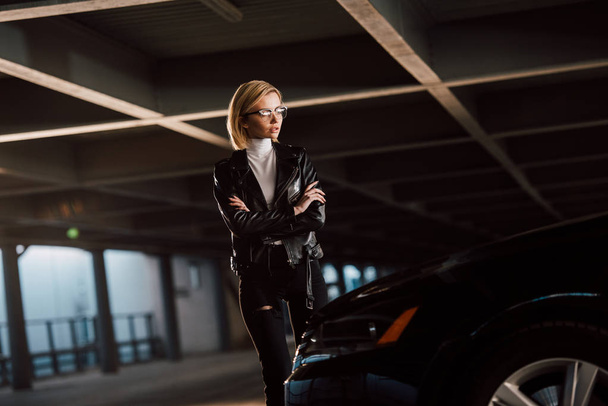 pretty young blonde woman in glasses standing with crossed arms near car  - Fotoğraf, Görsel
