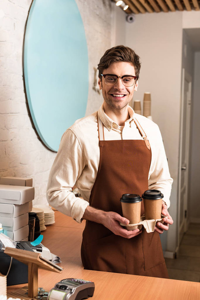 Barista sonriente en vasos sosteniendo portavasos para llevar con café
 - Foto, Imagen