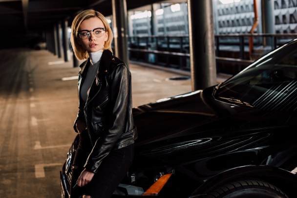 pensive young woman in glasses standing in parking near black car - Fotoğraf, Görsel