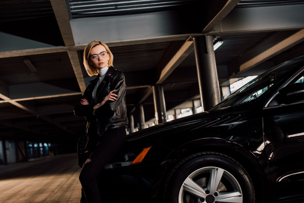 serious young woman in glasses standing in parking with crossed arms near black car - Fotografie, Obrázek