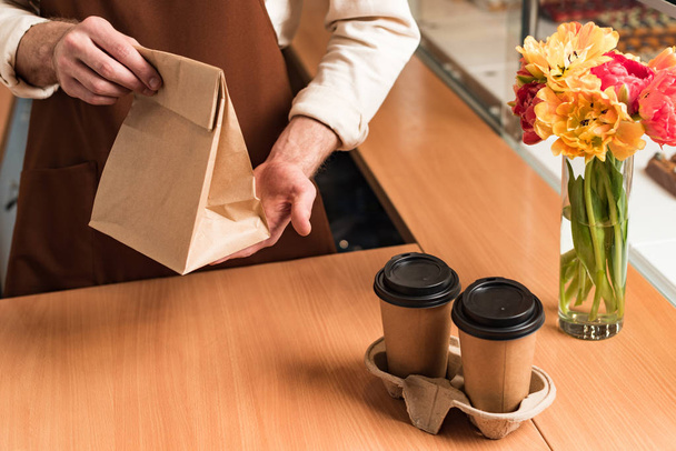Μερική θέα της μπάρμαν σε καφέ ποδιά με χάρτινη σακούλα και καφέ για να πάει - Φωτογραφία, εικόνα