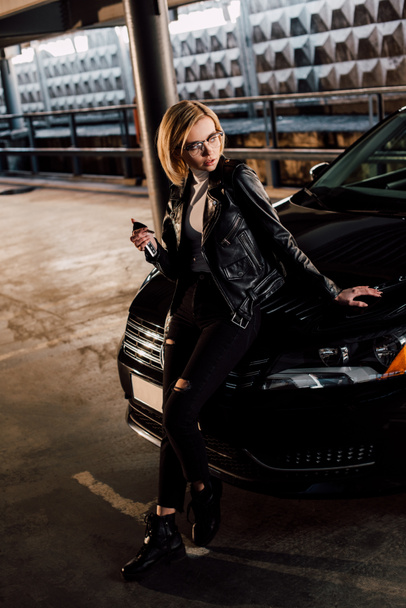 serious young woman in glasses standing with key in parking near black car - Photo, Image