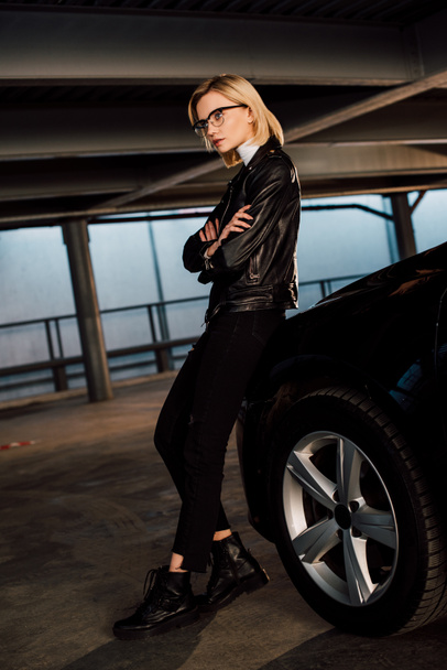 confident young woman in glasses standing in parking with crossed arms near black car - Foto, imagen