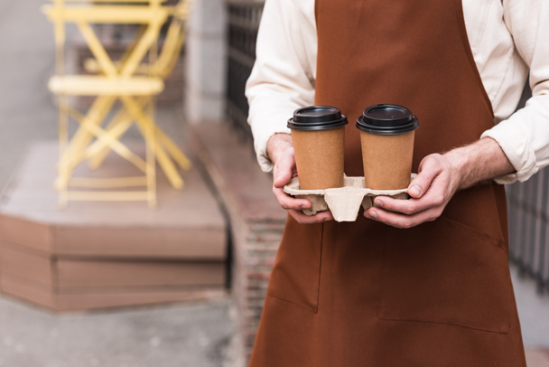 Обрезанный вид на бариста в коричневом фартуке, держащего носильщик чашки с кофе
 - Фото, изображение