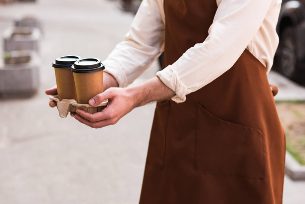 Cropped view of barista in brown apron holding take-out cup carrier with coffee - Foto, Imagen
