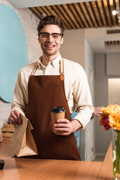 Smiling barista in glasses holding paper bag and disposable cup of coffee - Foto, imagen