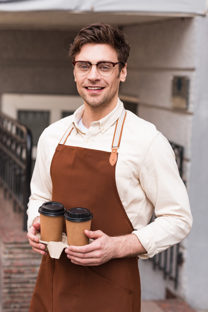 Smiling barista in glasses holding take-out cup carrier with coffee - Foto, imagen