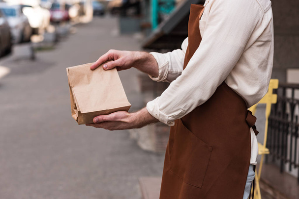 Вид на баристу в коричневом фартуке с бумажным пакетом на улице
 - Фото, изображение