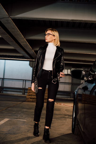 blonde woman in glasses holding key in parking near black automobile  - Foto, Imagen