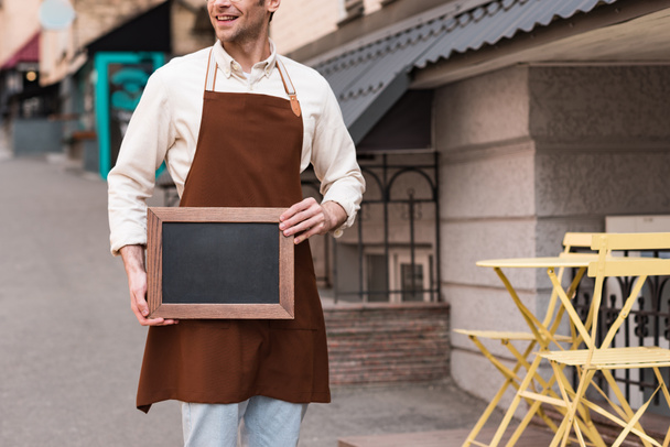 Cropped view of smiling barista in brown apron holding chalkboard menu on street - Photo, Image