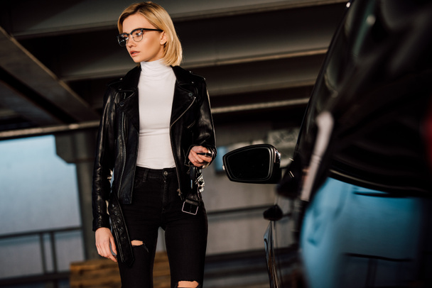 attractive girl in glasses holding key in parking near black automobile  - Zdjęcie, obraz