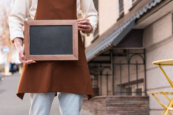 Cropped view of barista in brown apron holding chalkboard menu on street - Фото, изображение