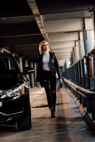 attractive blonde girl walking in parking with key near black automobile  - Foto, afbeelding
