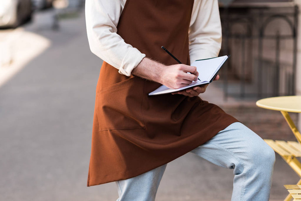 Cropped view of barista in brown apron writing in notebook on street - Foto, Imagen