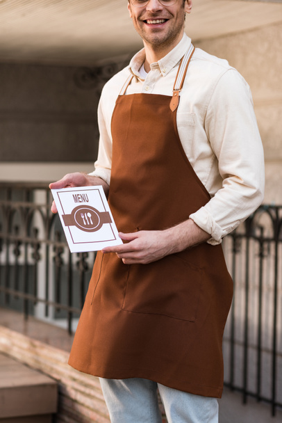 Vista cortada de sorrir barista em avental segurando menu na rua
 - Foto, Imagem