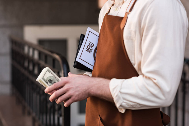 Partial view of barista in apron holding menu and money on street - Foto, Bild