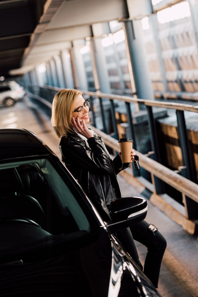 mosolygós szőke nő áll a parkolás eldobható csésze, miközben beszél okostelefon közelében fekete autó  - Fotó, kép