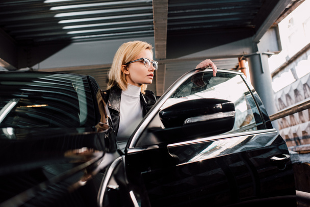 confident attractive girl in glasses near black automobile in parking  - Фото, изображение