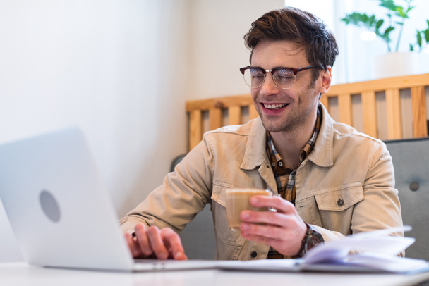 Smiling freelancer in glasses holding cup of coffee and typing on laptop keyboard - Foto, Bild