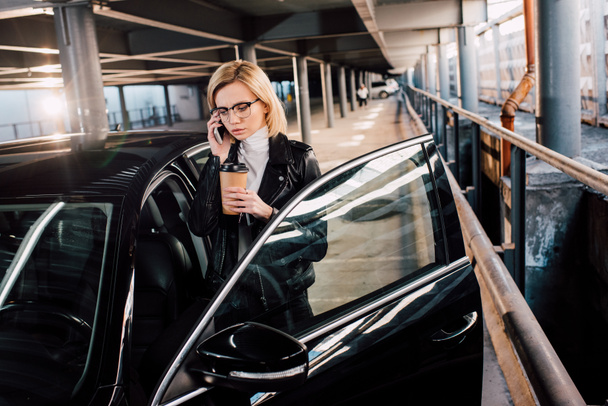 poważna dziewczyna w okularach rozmawiając na smartfonie i trzymając kawę, aby przejść w pobliżu czarnego samochodu  - Zdjęcie, obraz