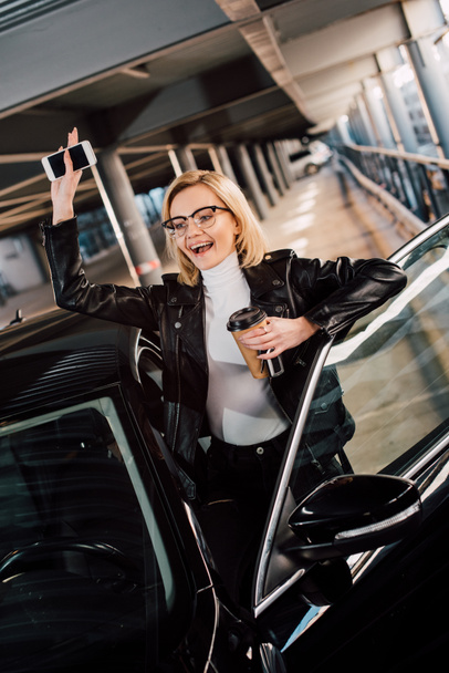happy girl holding smartphone and disposable cup and waving hand near car  - Photo, Image