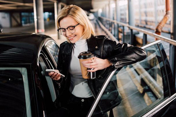 cheerful girl looking at smartphone and holding disposable cup near car  - Foto, Imagem