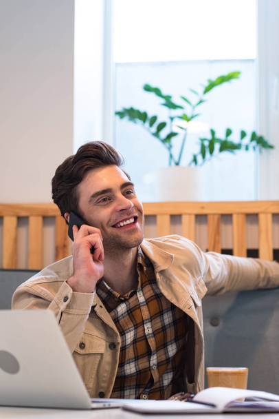Cheerful freelancer in jacket talking on smartphone in cafe - Foto, Bild