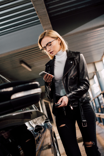 serious and attractive girl holding smartphone and key near black car  - Photo, Image