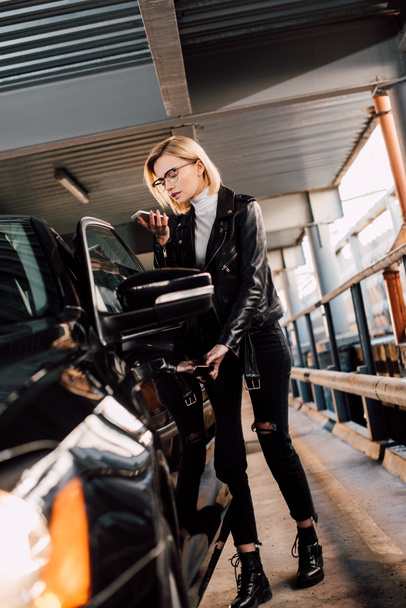 blonde attractive girl looking at smartphone while standing near car  - Zdjęcie, obraz