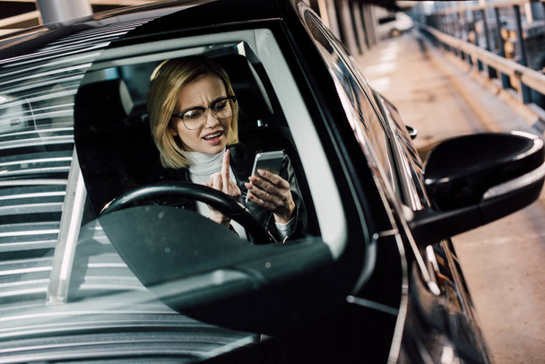 emocjonalna dziewczyna w okularach pokazując środkowy palec patrząc na smartphone w samochodzie  - Zdjęcie, obraz