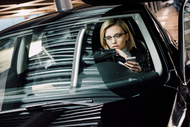 upset blonde girl in glasses holding smartphone in car  - Fotografie, Obrázek