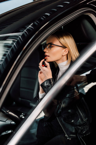 attractive blonde girl in glasses applying lipstick in car  - Φωτογραφία, εικόνα