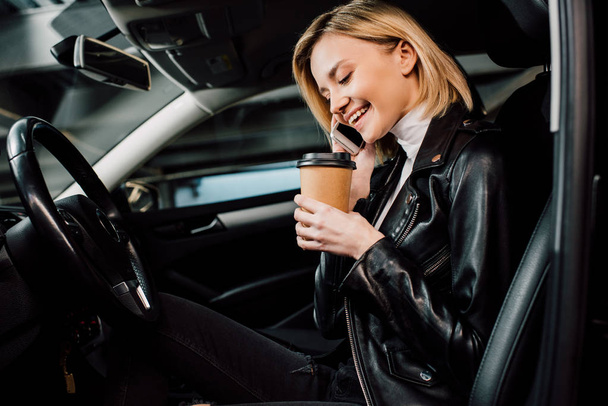 cheerful blonde girl holding paper cup and talking on smartphone in car  - Photo, Image