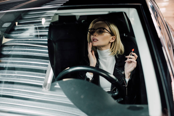bonita chica rubia en gafas sentado en el coche y la aplicación de lápiz labial
  - Foto, Imagen