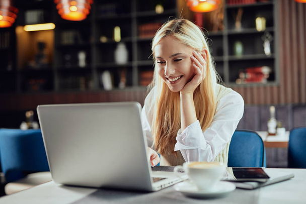 Piękna uśmiechnięta kaukaski blond kobieta blogger ubrany inteligentny casual siedzi w kawiarni i za pomocą laptopa. - Zdjęcie, obraz