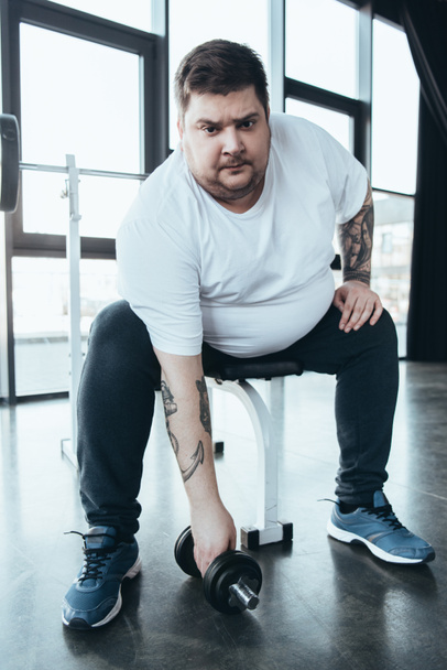 Hombre tatuado con sobrepeso mirando a la cámara y haciendo ejercicio con mancuerna en el centro deportivo
 - Foto, imagen