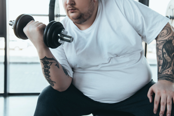 visão parcial do homem tatuado com excesso de peso no treinamento de camiseta branca com haltere no ginásio
 - Foto, Imagem
