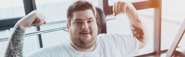 panoramic shot of overweight tattooed man smiling and showing muscles at gym with sunlight - Foto, Imagen