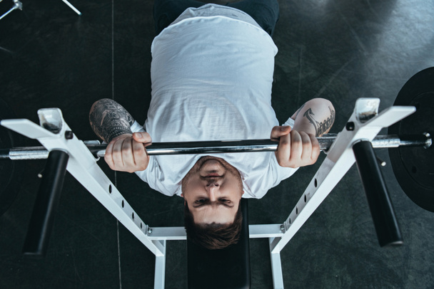vista superior del hombre tatuado con sobrepeso en entrenamiento de camiseta blanca con barra en el gimnasio
 - Foto, Imagen