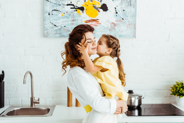 cute daughter kissing smiling beautiful mother in kitchen - Foto, afbeelding