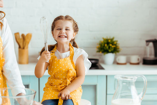 excited daughter in polka dot yellow apron holding balloon whisk next to mother in kitchen - Photo, Image