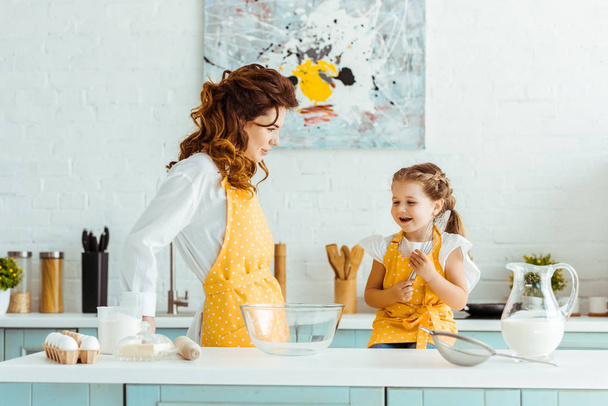mother in polka dot yellow apron looking at excited daughter in kitchen - Foto, imagen