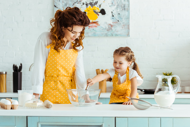 mother in polka dot yellow apron looking at cute daughter with balloon whisk in kitchen - Photo, Image