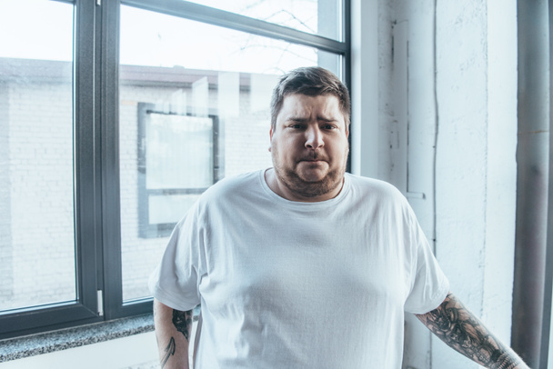 overweight tattooed man in white t-shirt looking at camera at gym - Fotó, kép