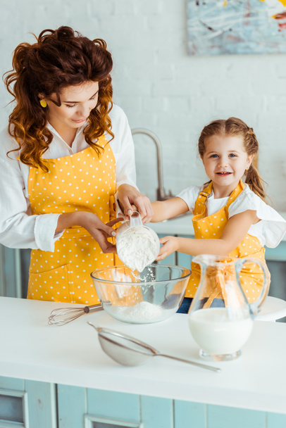 mother helping happy daughter adding flour to bowl in kitchen - Fotoğraf, Görsel