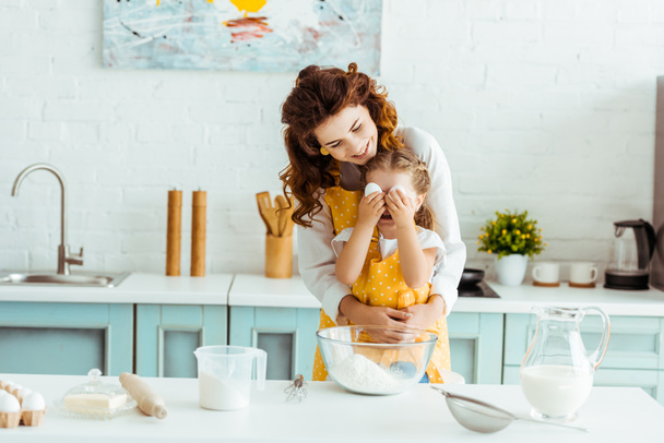 happy mother hugging daughter while kid holding eggs in front of eyes in kitchen - Photo, Image