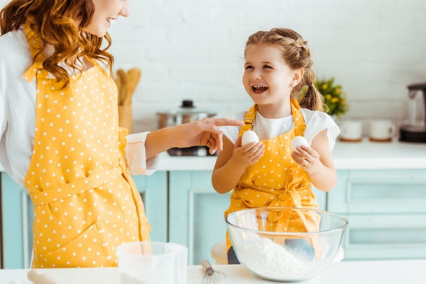 excited emotional daughter holding eggs while mother in yellow polka dot apron pointing with finger in kitchen - Фото, изображение