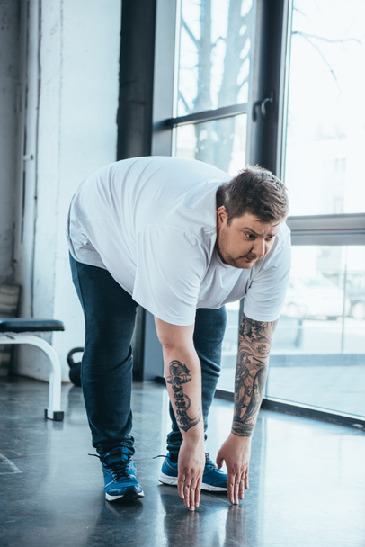 υπέρβαρο άντρας με τατουάζ σε λευκό μπλουζάκι κάνει τέντωμα άσκηση στο αθλητικό κέντρο - Φωτογραφία, εικόνα