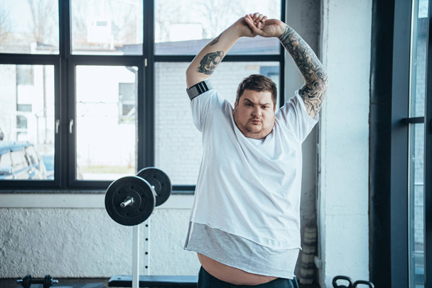 надмірна вага татуйованого чоловіка Дивлячись на камеру і роблячи вправи для розтягування в спортзалі
 - Фото, зображення