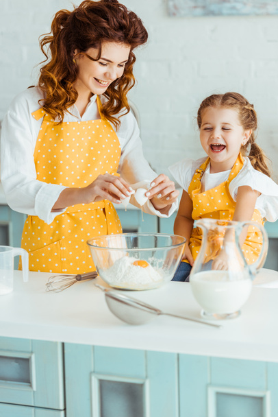 happy mother and daughter adding smashed egg to flour in bowl while cooking together in kitchen - Foto, Imagem